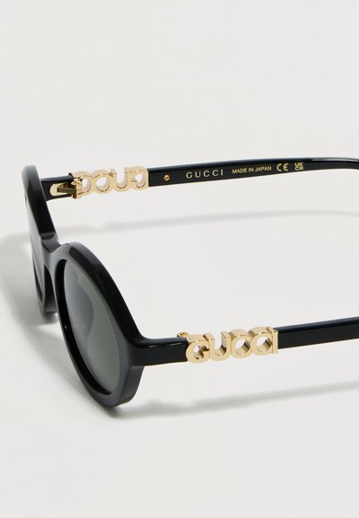 Μαύρα γυαλιά ηλίου Gucci με στρογγυλούς φακούς, χρυσό λογότυπο Gucci στα πλαινά και σήμανση "Κατασκευάστηκε στην Ιαπωνία" στο εσωτερικό του βραχίονα.