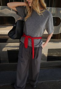 Maglione grigio a maniche corte con cintura rossa, abbinato a pantaloni grigi larghi e una borsa tote nera, ambientato su uno sfondo riflettente.