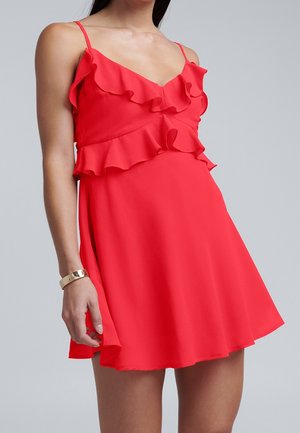 Woman wearing a sleeveless bright red dress with ruffles along the neckline and waist, standing against a plain gray background.