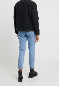 Svart denimjacka med pälskrage, ljusblå tapered jeans och svarta läderankelstövlar med siddragkedja. Bakifrån.