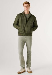 Chaqueta bomber verde oliva con cierre de cremallera frontal, tela suave, puños y dobladillo acanalados, combinada con jeans verde claro y zapatillas beige.