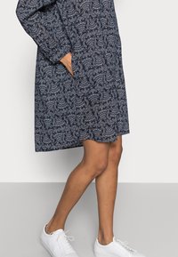 Robe navy avec un motif paisley blanc, manches longues, poches latérales et coupe décontractée. Associée à des baskets blanches pour un look décontracté.