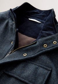 Veste en chevron bleu marine avec fermeture éclair dorée et boutons-pression, montrant une doublure de col en velours côtelé doux et une poche avant à rabat.