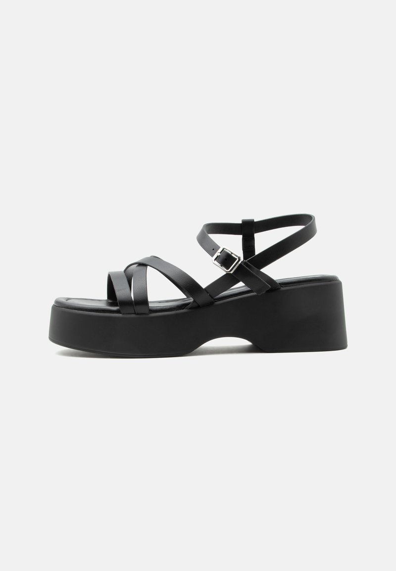 Sandale à plateforme noire avec des brides croisées, une boucle à la cheville et un talon épais sur fond blanc.