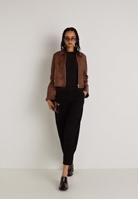 Smeđa kožnata skraćena jakna preko crnog topa, uparena s crnim hlačama i tamnim, sjajnim čizmama do gležnja. Dodaci uključuju sunčane naočale i telefon.