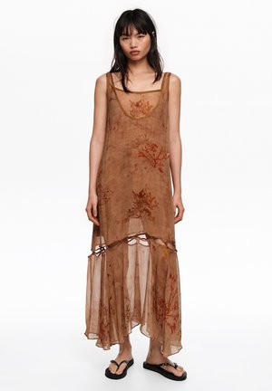 Kobieta z prostymi czarnymi włosami, ubrana w bezrękawnikową, przezroczystą, maxi sukienkę w brązowe kwiaty oraz czarne sandały, stojąca na białym tle.