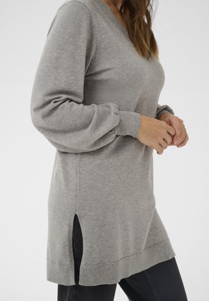 Egy nő szürke, hosszú ujjú kötött pulóvert visel oldalsó hasítékkal és fekete nadrággal, kezei finoman összekulcsolva a dereka előtt.