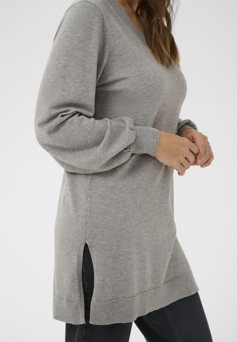 Egy nő szürke, hosszú ujjú kötött pulóvert visel oldalsó hasítékkal és fekete nadrággal, kezei finoman összekulcsolva a dereka előtt.