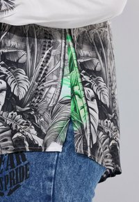 Gros plan sur une chemise à motif de feuilles tropicales en noir et blanc avec une touche de vert, portée sur un jean bleu avec un imprimé de texte noir.