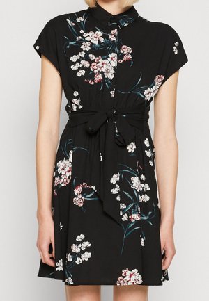 Mujer vistiendo un vestido negro de manga corta con patrones florales blancos y rosas y un cinturón atado a la cintura.