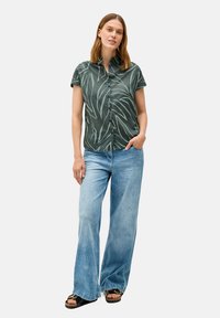 Camicetta verde a maniche corte con motivi di foglie, chiusura a bottoni, abbinata a jeans ampi blu chiaro e sandali neri con dettagli dorati.
