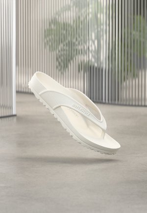 Balta Birkenstock EVA įlinkio tipo sandalė, sklandanti virš pilkos grindų dangos, fone matomas neryškus augalas ir vertikalios plokštės.