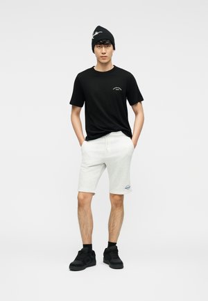 Joven con gorro negro, camiseta negra, pantalones cortos gris claro, calcetines negros y zapatillas negras, de pie con las manos en los bolsillos.