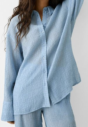 Woman wearing textured light blue button-up long-sleeve shirt and matching pants standing against a plain background.