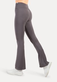 Szare, rozszerzane spodnie do jogi wykonane z gładkiego, elastycznego materiału. Posiadają wysoki pas i detal w postaci bocznego rozcięcia, zestawione z białymi butami sportowymi.