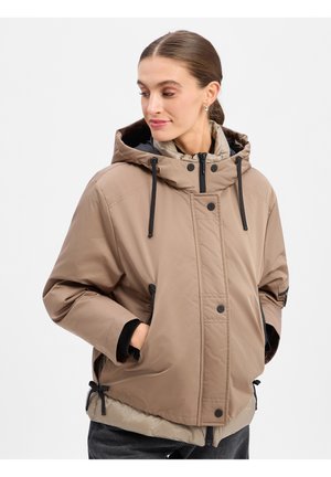 Frau mit zurückgebundenem Haar, die eine beige Kapuzenjacke mit schwarzen Kordeln trägt, die Hände in den Taschen, schaut nach links unten.
