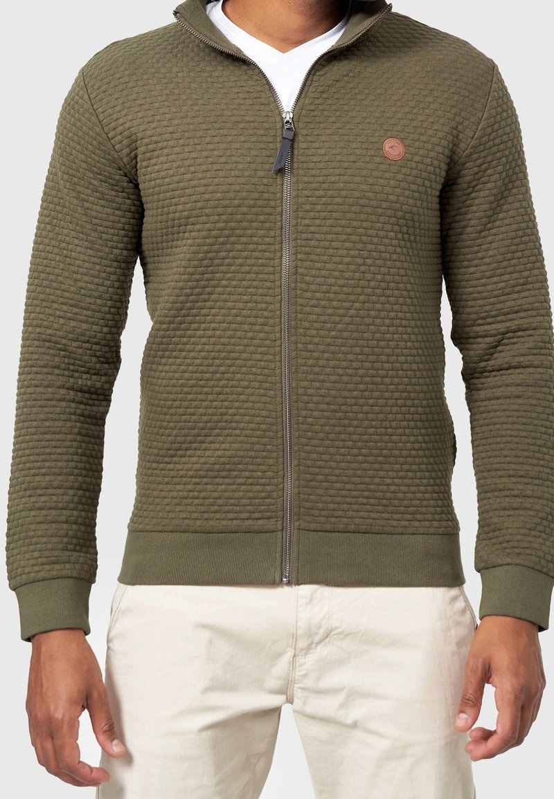Homme portant une veste zippée texturée vert olive par-dessus une chemise blanche et un pantalon beige, debout bras détendus.