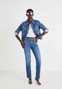 Denim jakna v srednje modri barvi z dvema žepoma na prsih, v kombinaciji z belim skrajšanim vrhom in oprijetimi modrimi jeans hlačami, poudarjena s sandali na vezalke.