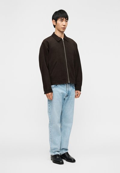 Άνδρας στέκεται με πρόσωπο προς τα εμπρός, φοράει σκούρο καφέ μπουφάν με φερμουάρ, ανοιχτόχρωμα μπλε τζιν και μαύρα επίσημα παπούτσια μπροστά σε απλό λευκό φόντο.