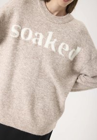 Femme portant un pull en laine beige avec "Soaked" en grandes lettres blanches sur la poitrine, associé à un pantalon noir.