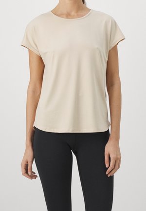 Femme portant un t-shirt beige uni à manches courtes et un pantalon noir ajusté, debout devant un fond neutre.