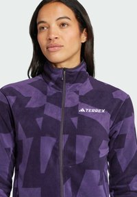 Chaqueta de forro polar en púrpura con un patrón geométrico abstracto. Presenta un cuello alto y una cremallera frontal completa. Logo visible en el lado izquierdo del pecho.