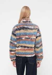 Chaqueta difusa y multicolor con rayas en azul, marrón, crema y negro. Cuello alto y puños elásticos; textura suave.