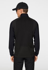 Giacca sportiva nera con colletto alto, maniche lunghe e retro texturizzato che presenta un motivo a rete e dettagli di branding discreti.