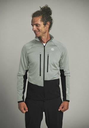 Mann mit lockigem Haar in grauem und schwarzem Reißverschluss-Pullover, schaut nach links, steht vor schlichtem grauem Hintergrund.