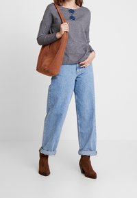 Haut gris à manches longues, jean bleu taille haute, bottines en daim marron, et un sac à bandoulière marron. Des lunettes de soleil rondes complètent le look.