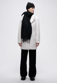 Bijeli kaput od umjetnog krzna, crna pletena kapa i šal. Crne široke hlače. Glatka tekstura s istaknutim detaljima resica na šalu.