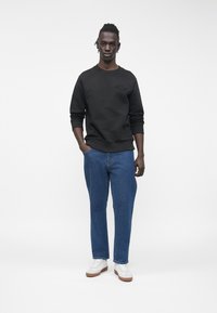 Mladý muž s copánky, oblečený v černé mikině, modrých džínách a bílých teniskách, stojící s jednou rukou v kapse na jednobarevném pozadí.