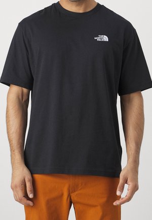 Mężczyzna w czarnej koszulce z krótkim rękawem z małym białym logo The North Face na lewej piersi oraz w pomarańczowych spodniach.