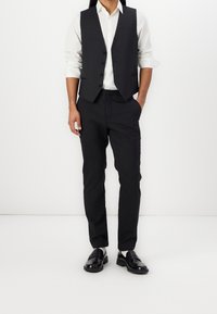 Schwarze Weste und Hose aus glattem Stoff, kombiniert mit einem weißen Hemd mit Knöpfen. Schwarze Schuhe runden den Look ab. Schlichtes und maßgeschneidertes Design.