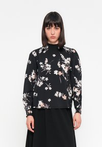 Donna con capelli scuri e lisci che indossa una blusa nera a maniche lunghe con fiori e collo alto, abbinata a una gonna nera, il tutto su uno sfondo bianco.