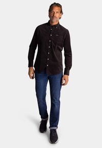 Zwarte corduroy overhemd met lange mouwen, met knoopsluiting kraag, borstzak en witte knopen, gecombineerd met donkerblauwe jeans en donkere sneakers.