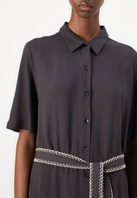 Robe noire à boutons avec manches courtes, en tissu texturé et ceinture crème contrastante avec couture en zigzag à la taille.