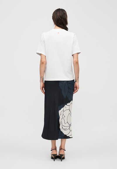 Γυναίκα με σκούρα μαλλιά φοράει λευκό μπλουζάκι με κοντά μανίκια και μαύρη midi φούστα με μεγάλο λευκό και σκουρομπλε λουλουδάτο μοτίβο, στέκεται γυρισμένη από την πλάτη.