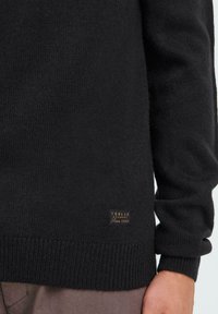 Black knitted sweater with ribbed cuffs and hem. Features a small brown brand tag on the lower side, showing texture and a simple design.