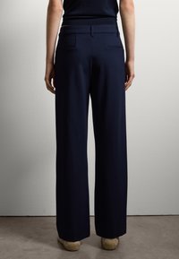 Pantaloni a gamba larga in tessuto blu navy, con due tasche posteriori e una texture liscia. Il cinturino è rifinito in modo elegante.