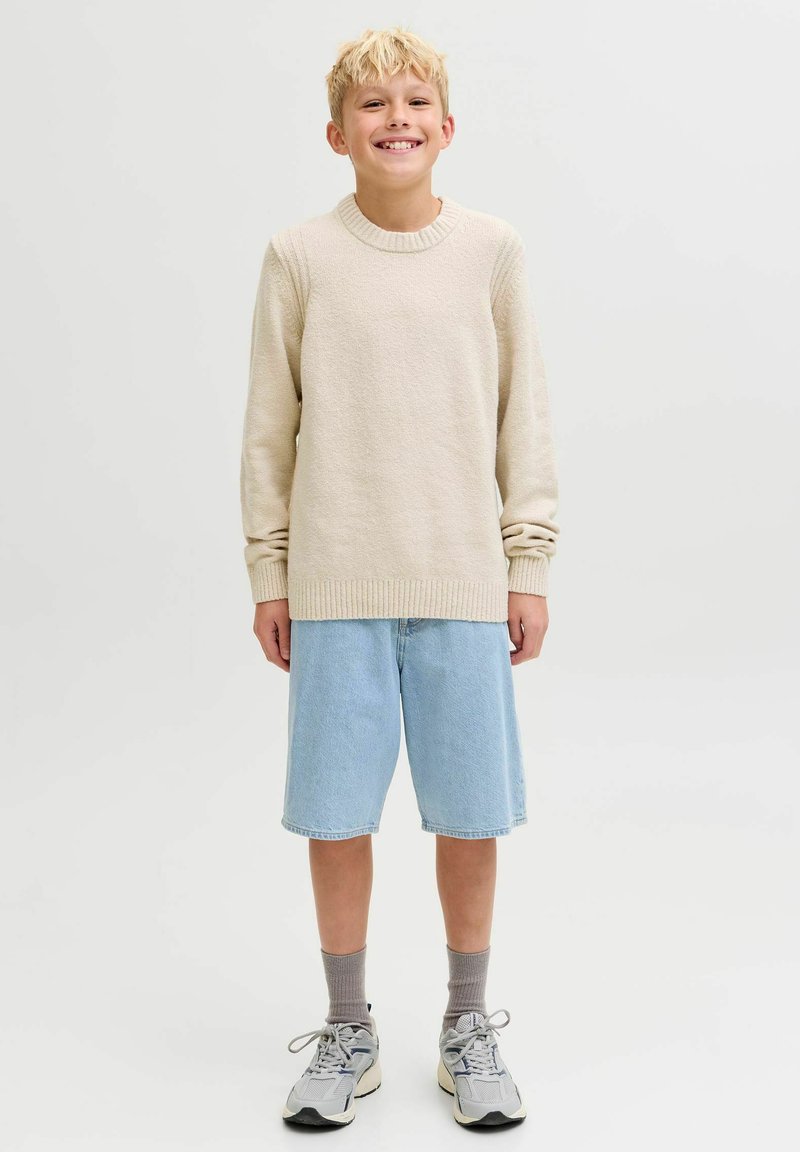 Šviesiai smėlinės spalvos megztinis su raukšlėtu apykaklėje, derinamas su šviesiai mėlynomis džinsinėmis šortais. Nešiojamas su pilkomis kojinėmis ir pilkais sportbačiais.