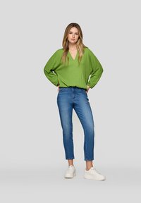 Frau steht mit den Händen in den Taschen, trägt eine grüne Langarm-Bluse mit V-Ausschnitt, blaue Jeans und weiße Sneakers vor einem schlichten Hintergrund.