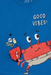 Blå bomulls t-skjorte med en tegneseriekrabbe i rødt, med teksten "GOOD VIBES!" Inkluderer illustrasjoner av en ball, iskrem og solbriller.