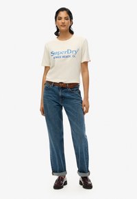 Cremefarvet t-shirt med blå "SuperDry" logo og tekst "Venice Beach, CA", parret med blå jeans og brun flettet bælte; læder loafers.