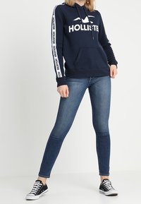 Felpa con cappuccio blu navy con logo bianco "HOLLISTER" e strisce laterali, abbinata a jeans skinny blu e sneakers nere.