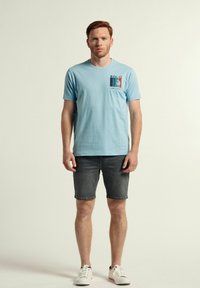 Camiseta de algodón de color azul claro con cuello redondo, con un estampado gráfico en el lado izquierdo, combinada con pantalones cortos de mezclilla oscura y zapatillas blancas.