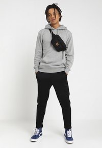 Felpa grigia con tasca frontale, marsupio nero, pantaloni neri attillati e sneakers blu con dettagli bianchi. Stile streetwear informale.