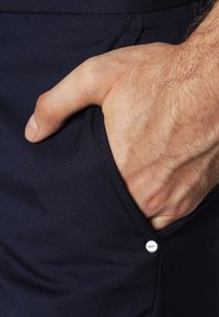 Námořnické modré kalhoty s texturovaným povrchem. Na přední straně je ruka v kapsi a na boku je stříbrný akcentovaný knoflík.