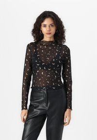 Schwarzes, transparentes Langarmoberteil mit einem Muster aus Monden und Punkten. Wellig geschnittener Saum und Kragen in Mockneck-Design. Kombiniert mit schwarzen Lederhosen.