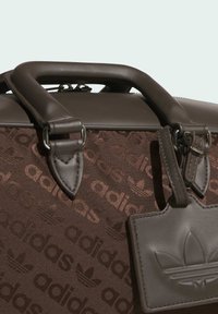 Brown sports bag with embossed Adidas logo pattern, leather handles, and a textured tag. Features a structured shape and metal hardware.